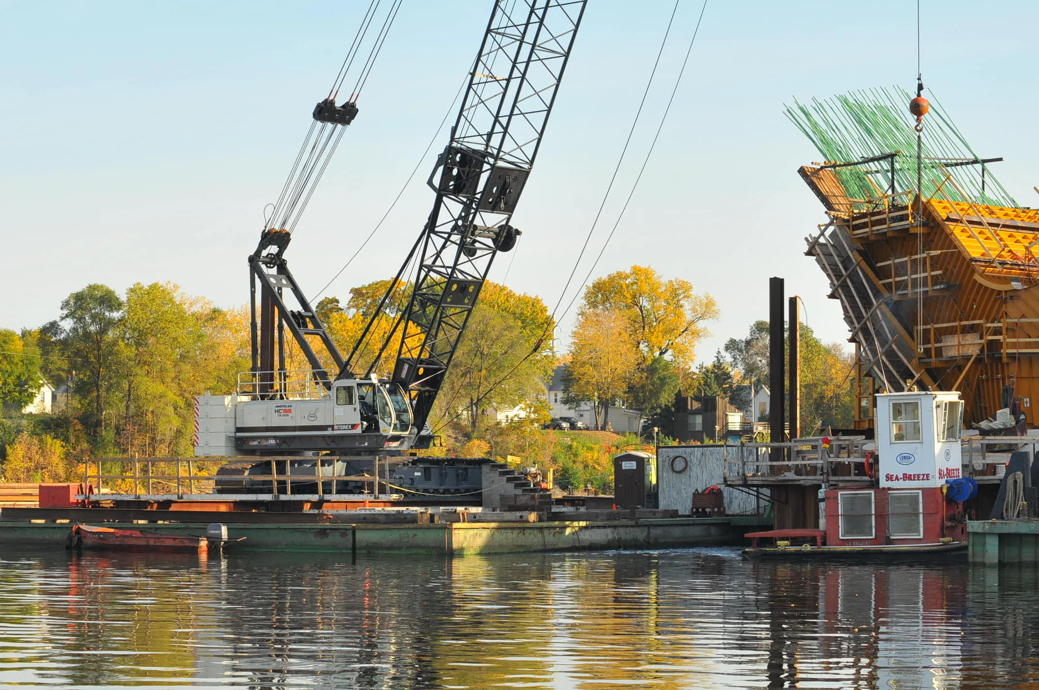 Construction crane on a barge building a bridge pier over water, surrounded by autumn trees.