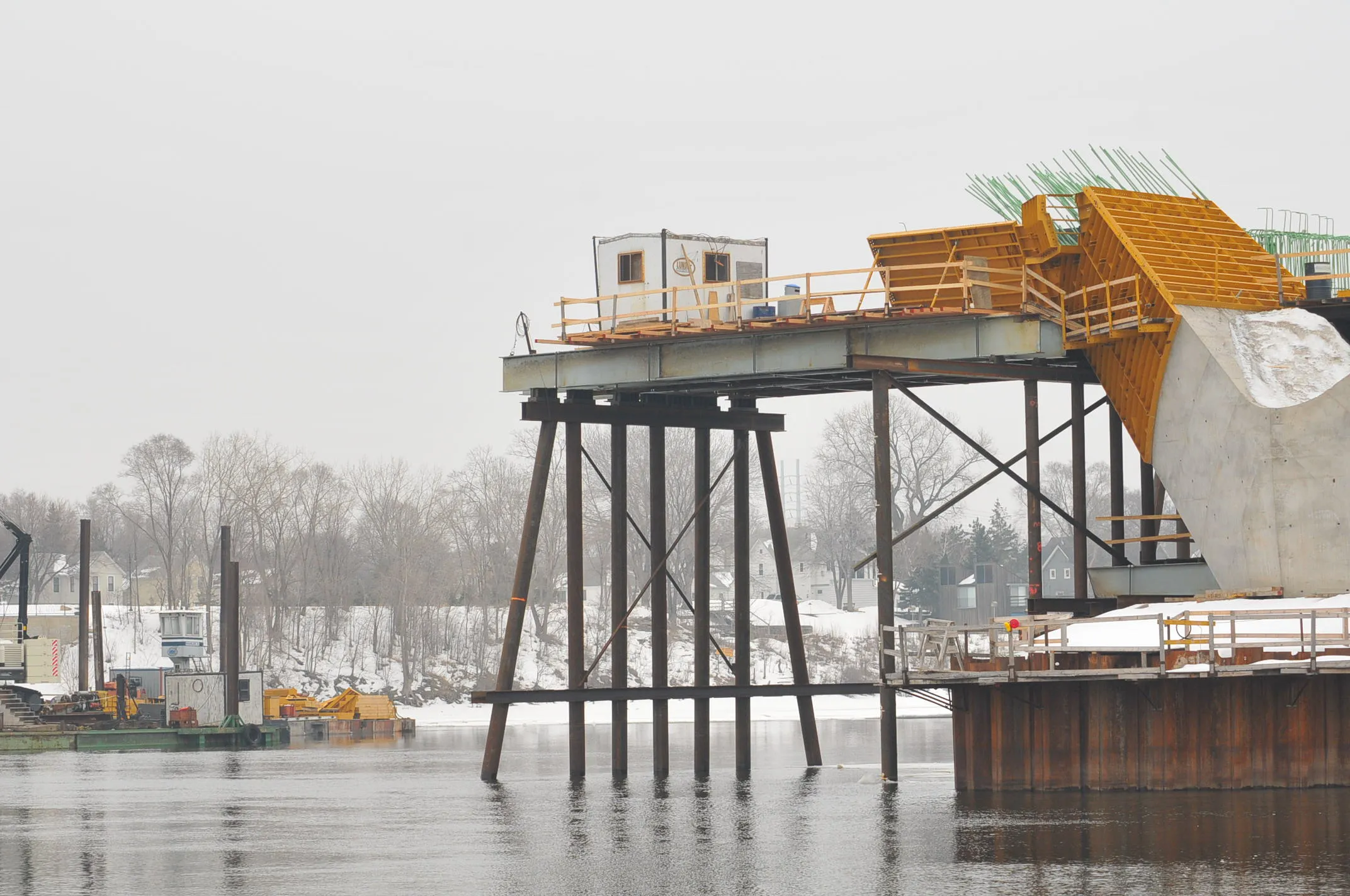 Bridge under construction over a snowy river, with concrete formwork, rebar, and temporary work sheds on site.