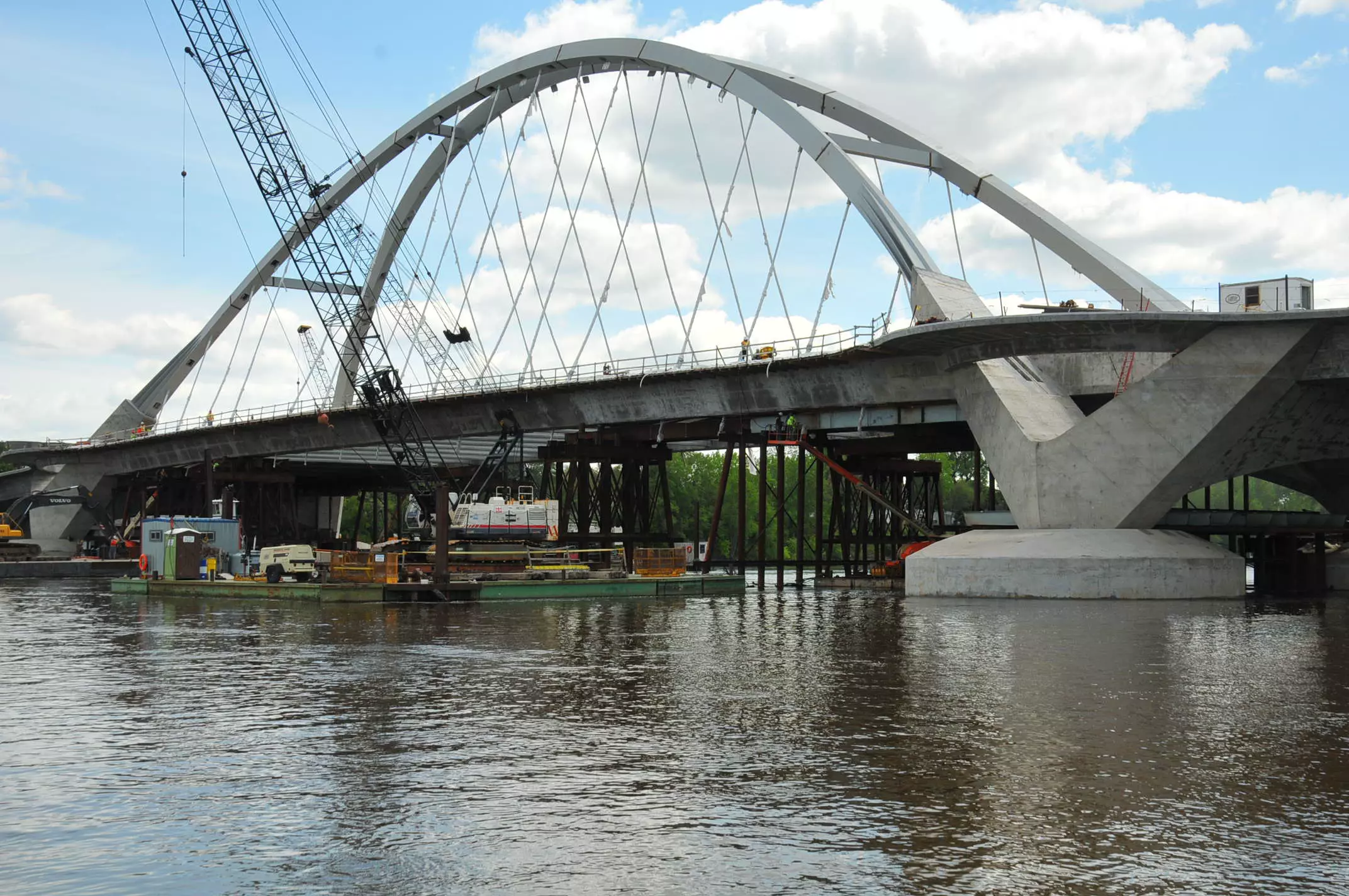 Arch bridge under construction over a river, with cranes, barges, and workers on the bridge under a cloudy sky.