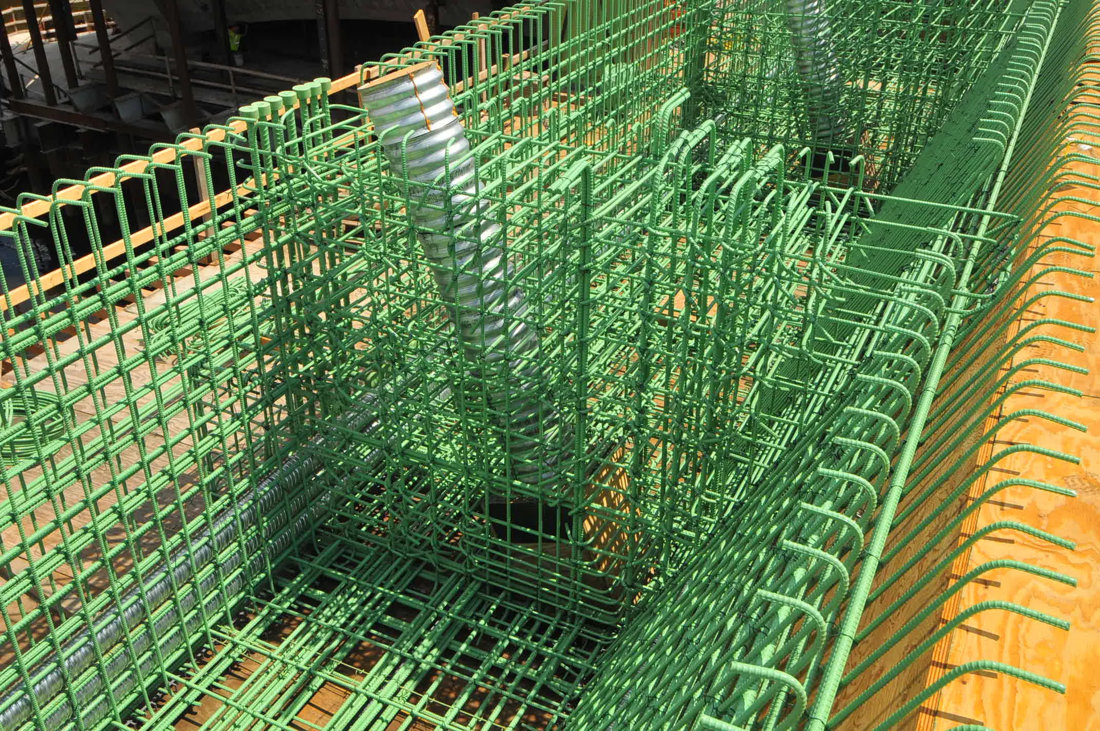 Green epoxy-coated rebar cage with corrugated metal pipes forming a concrete structure on a construction site.