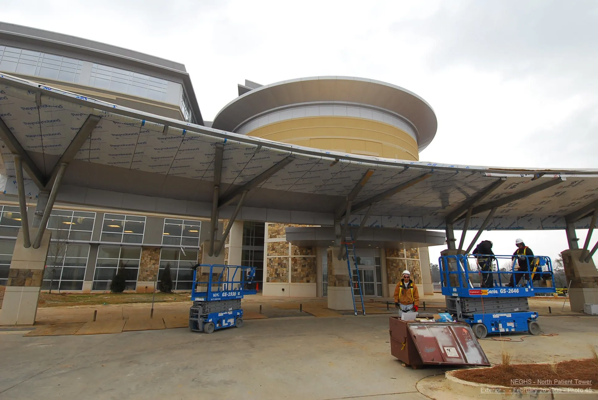 Hospital patient tower under construction. Modern entrance with large curved canopy, workers on scissor lifts.
