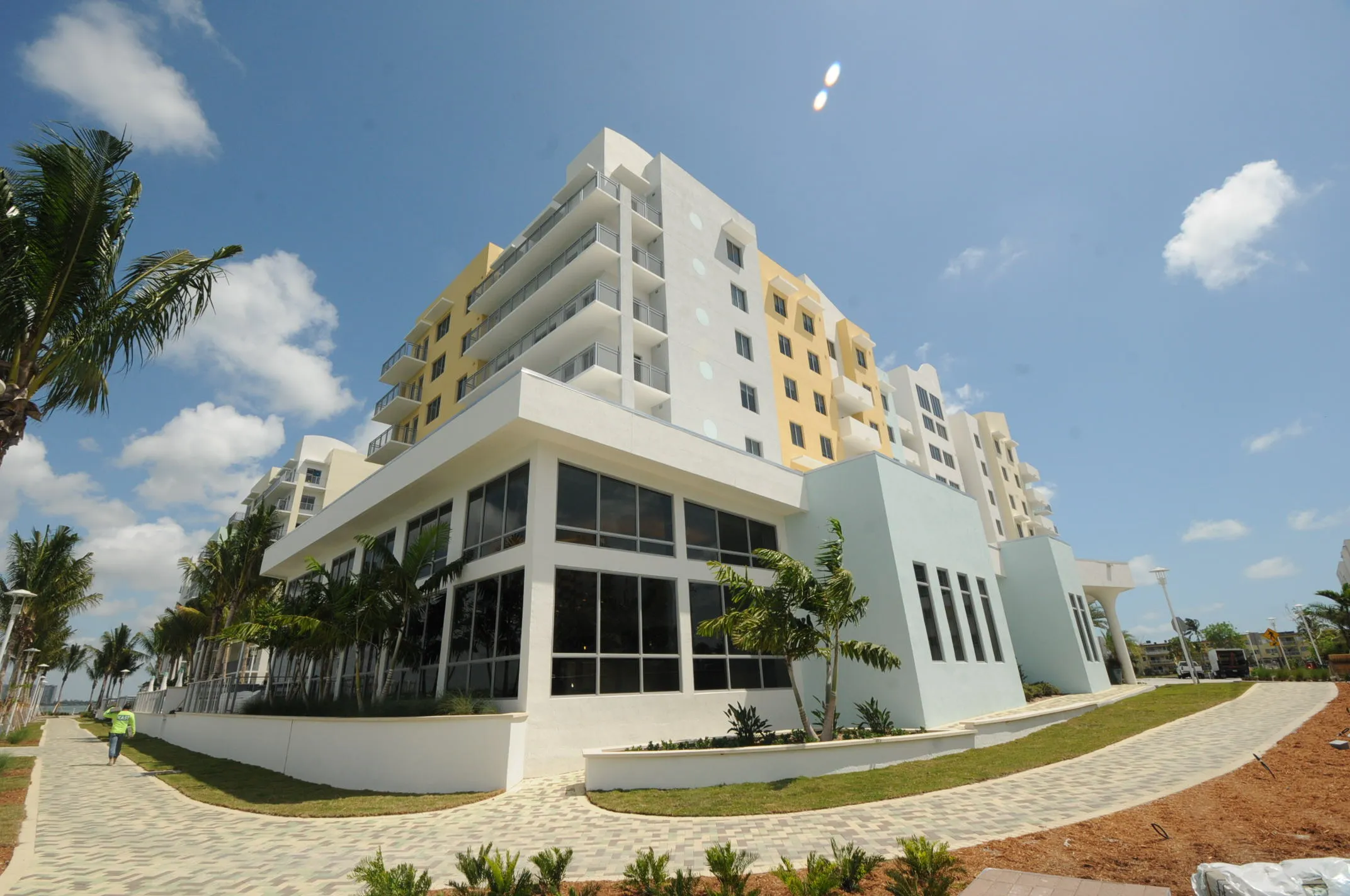 Large modern residential building with yellow and white sections, balconies, palm trees, and a person walking on a path.