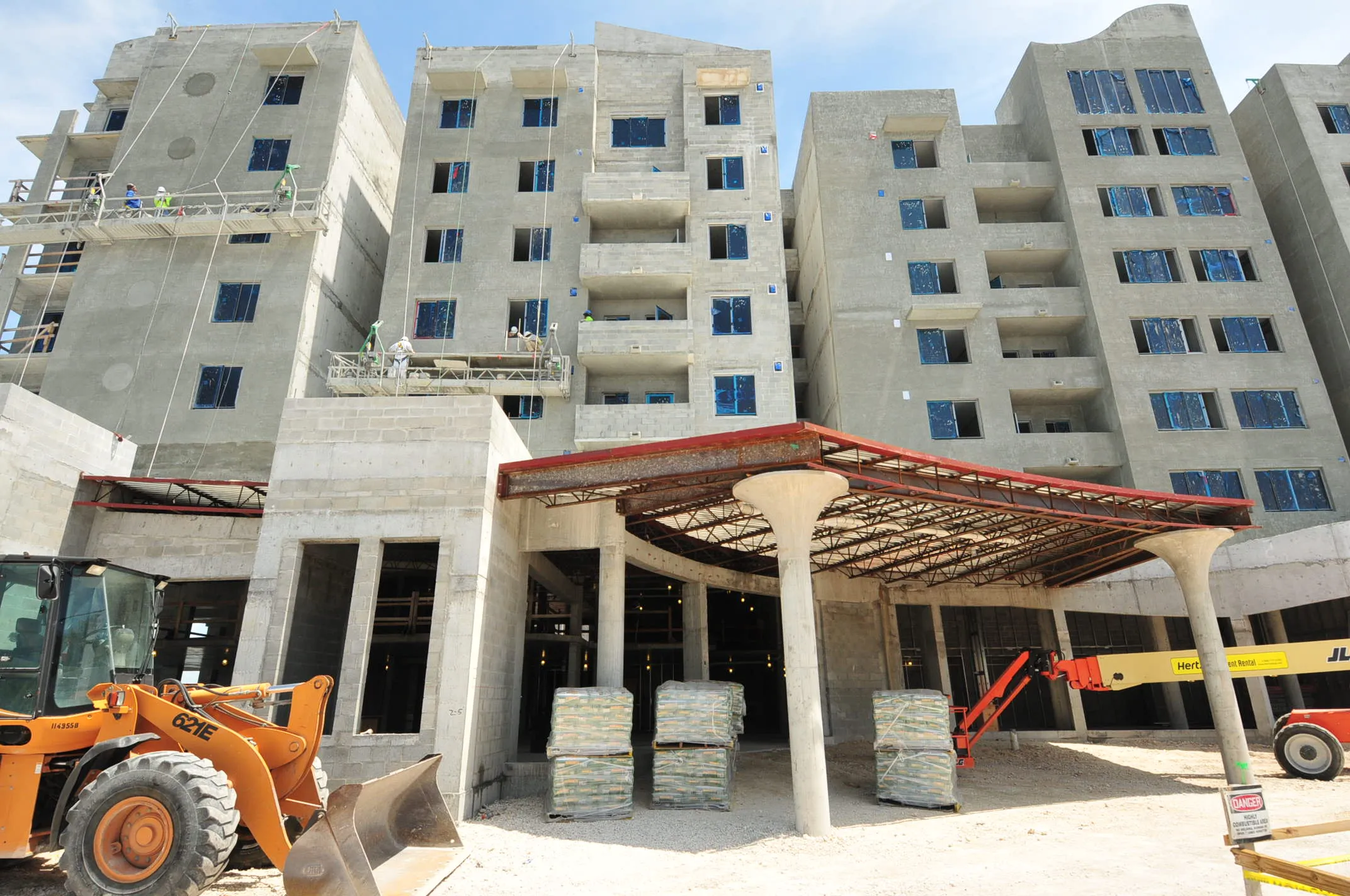 Multi-story buildings under construction. Workers on scaffolding, front-end loader, and boom lift visible.