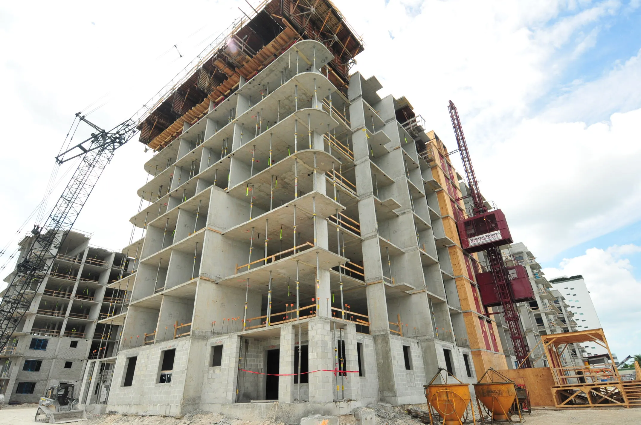 Concrete multi-story building under construction with two towering cranes against a cloudy sky.