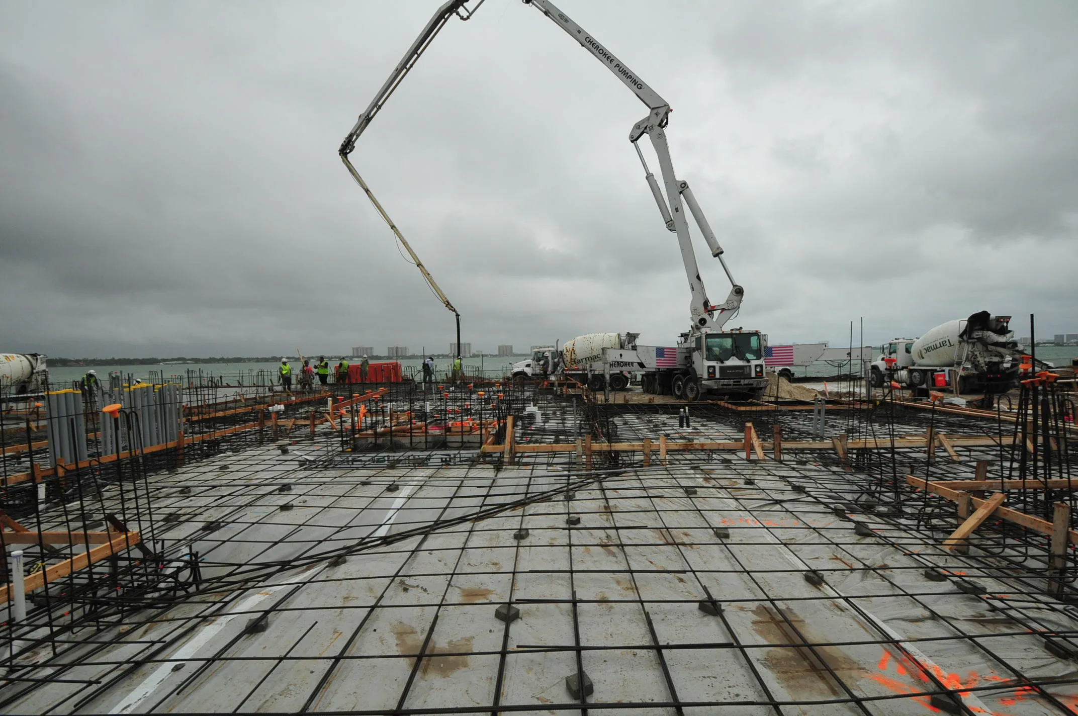 Construction workers oversee a concrete pump truck extending its boom over a rebar grid on a cloudy day by the bay.