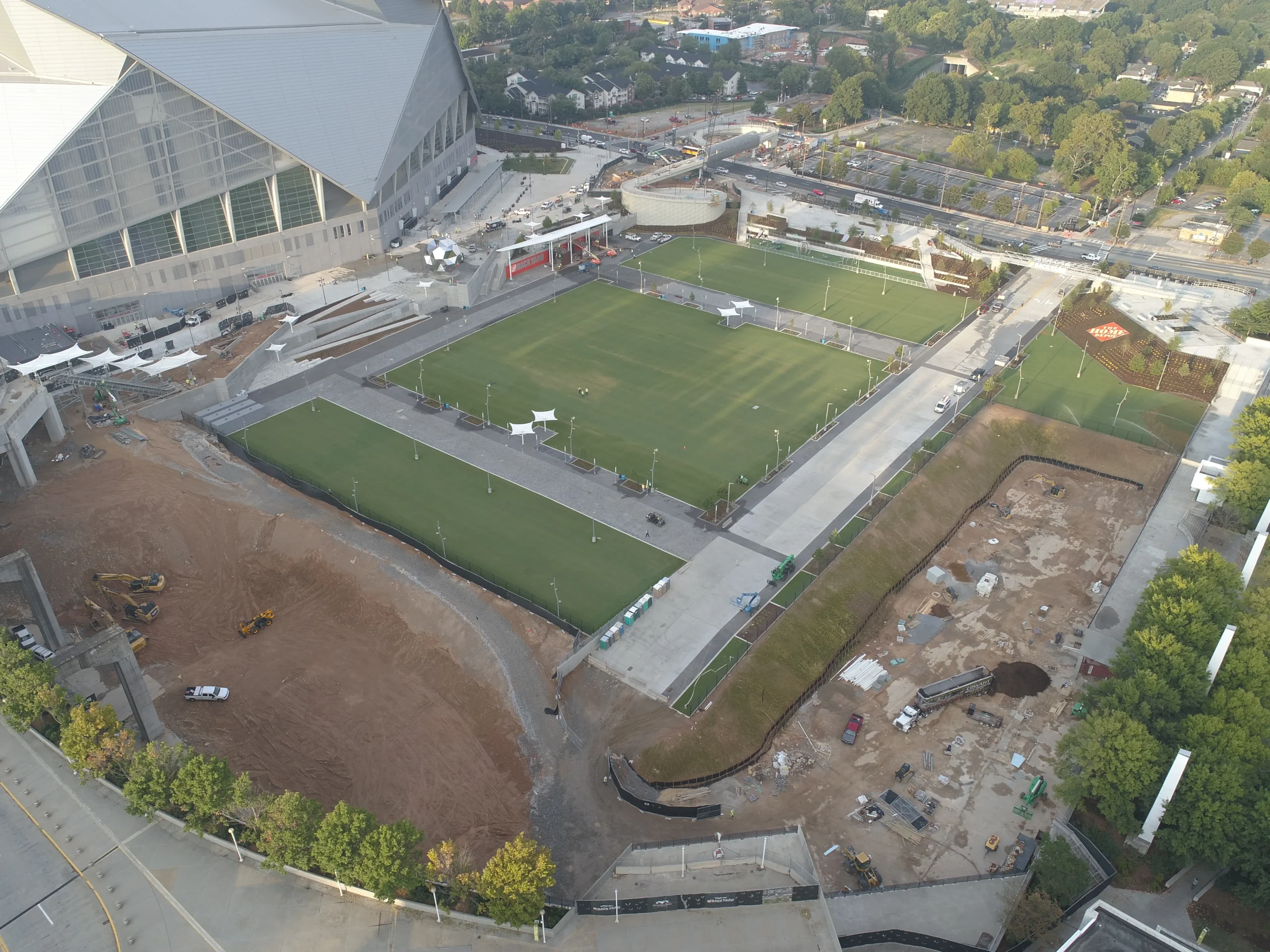 Aerial view of a modern stadium, large green fields, plazas, and surrounding construction sites.