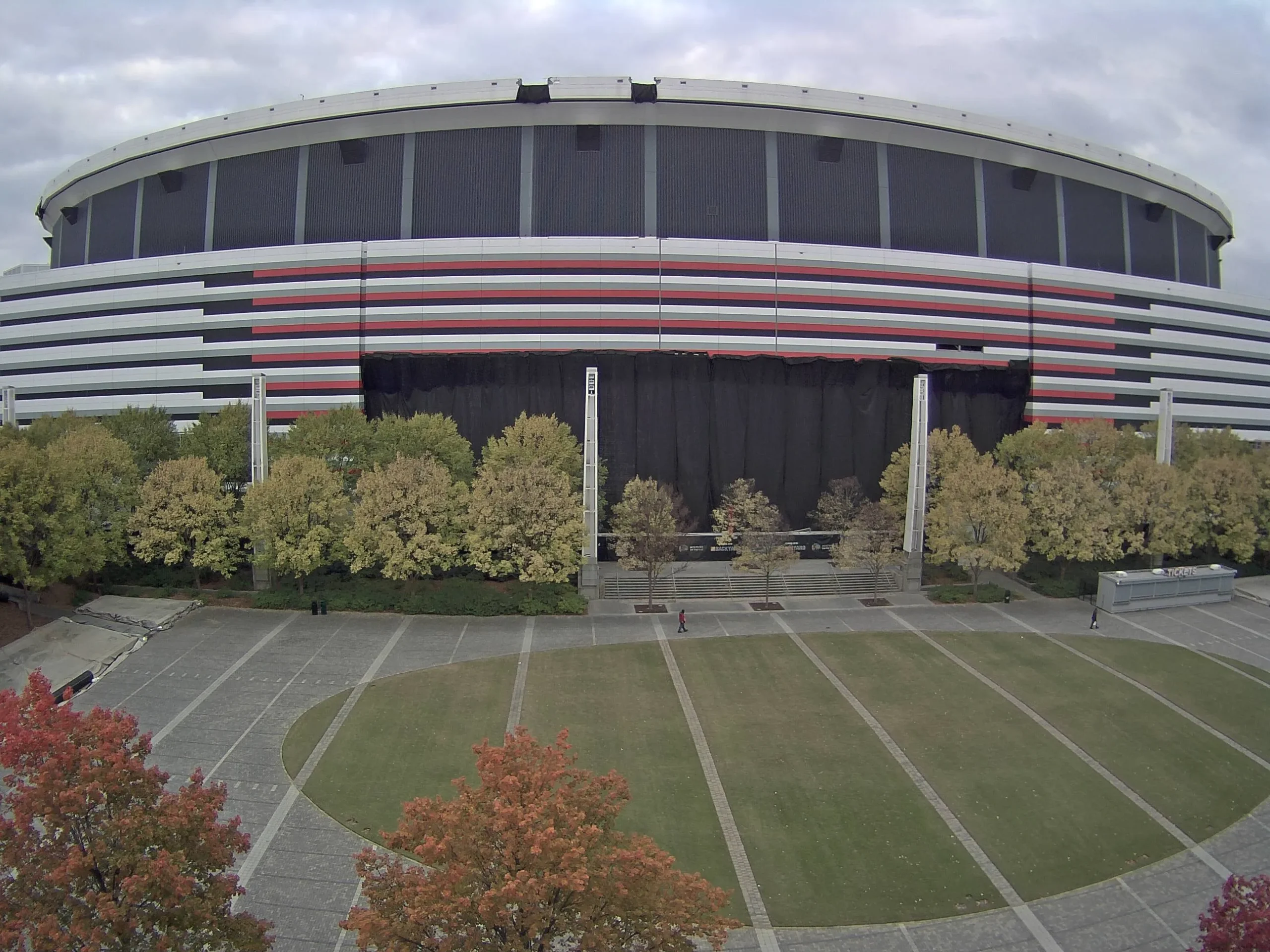 Large stadium with striped facade (white, black, red, gray) overlooking a plaza with grass and autumn trees.