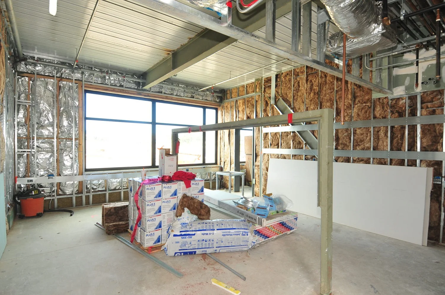 Interior of a building under construction showing exposed metal framing, insulation, a large window, and building materials.