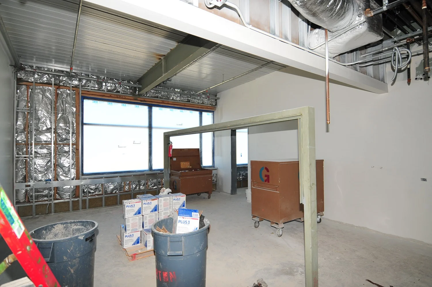 Commercial interior under construction, featuring exposed insulation, large windows, ceiling ducts, and building supplies.