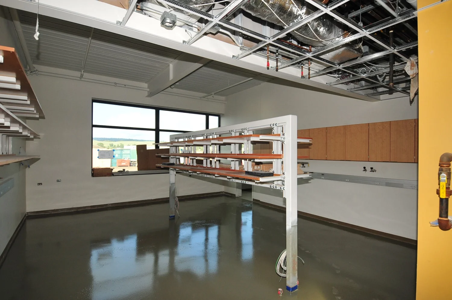 Empty lab or classroom under construction with exposed ceiling, wet floor, shelving, and large window.