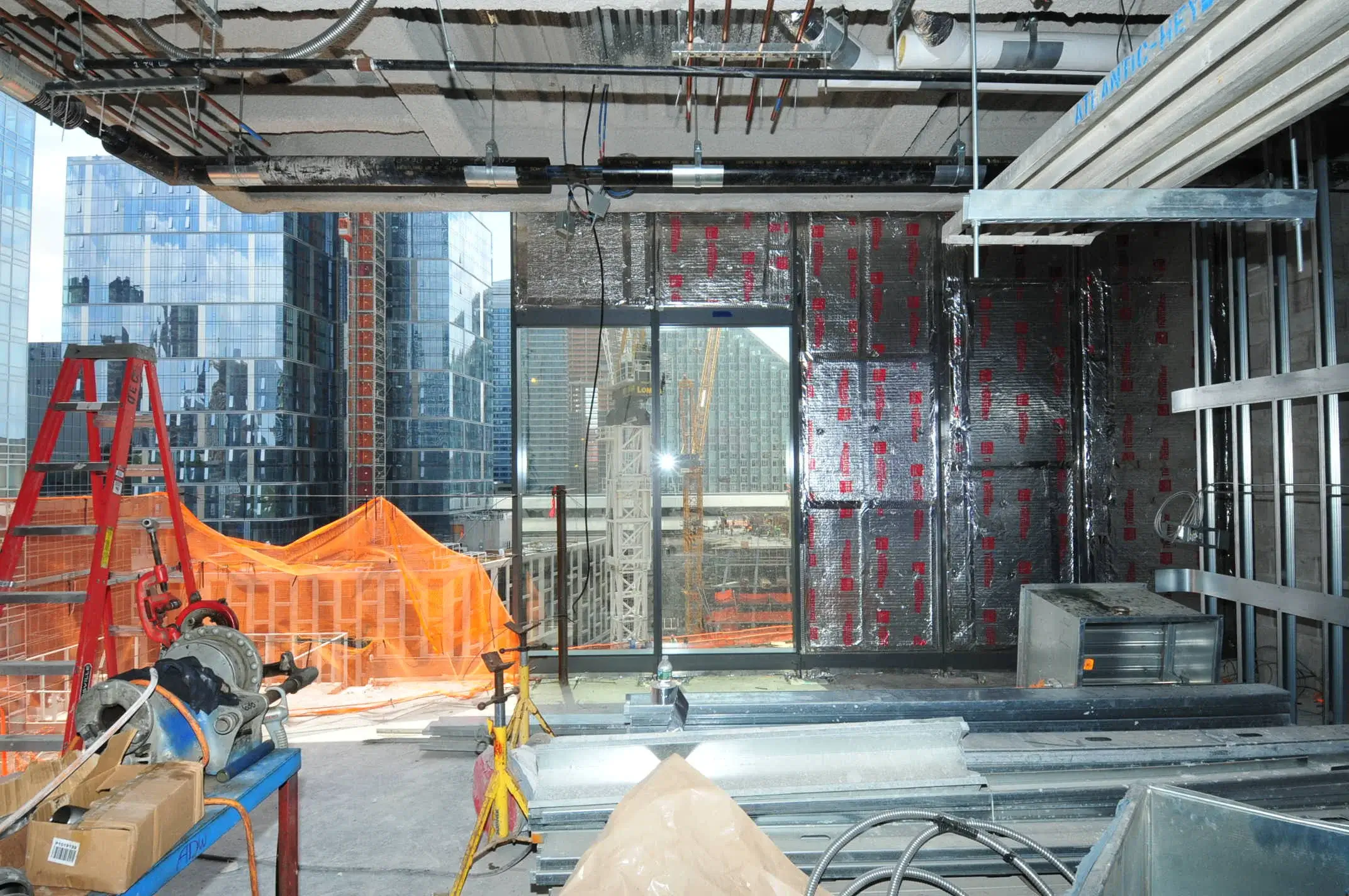 Indoor construction site with exposed pipes, tools, building materials, and a city view through large windows.