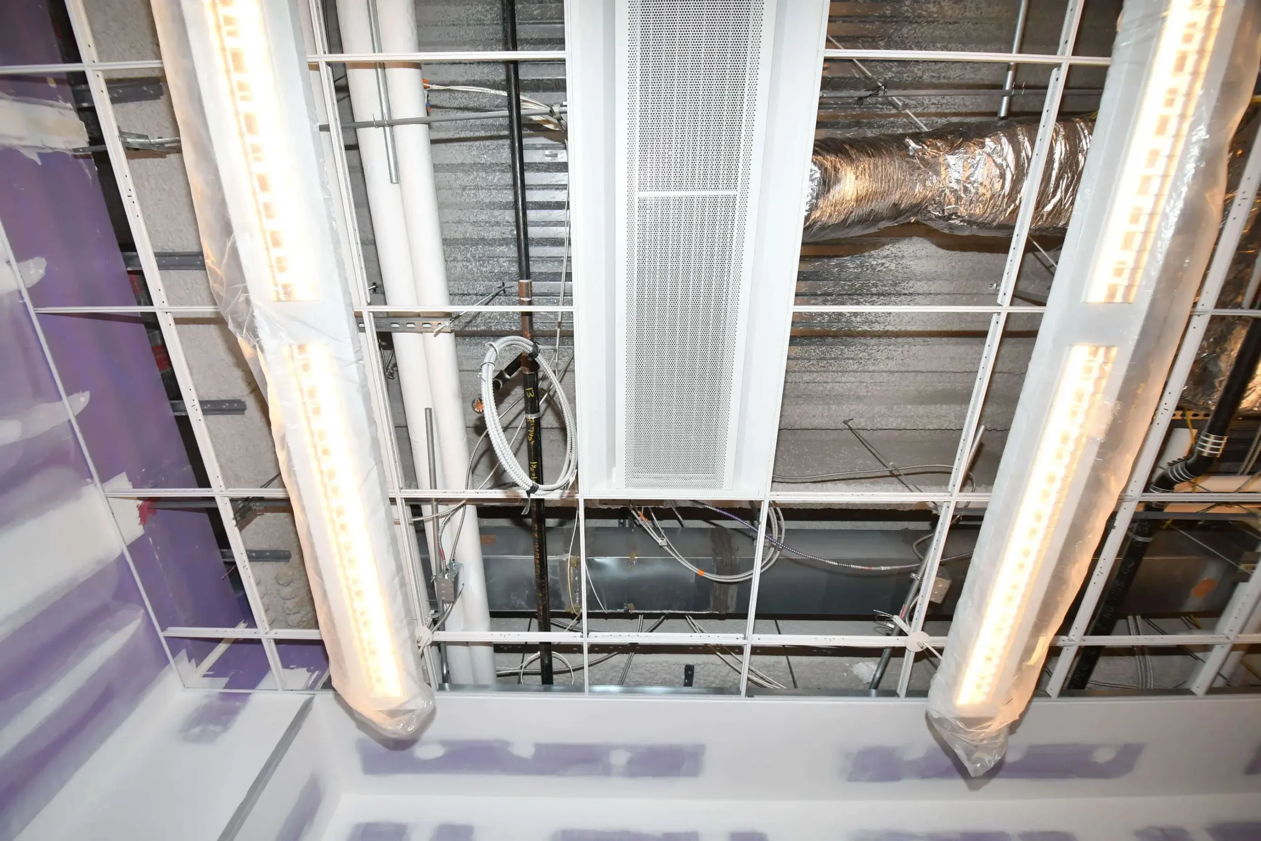 Construction site ceiling with exposed grid, new lights in plastic, ductwork, pipes, and unpainted drywall.