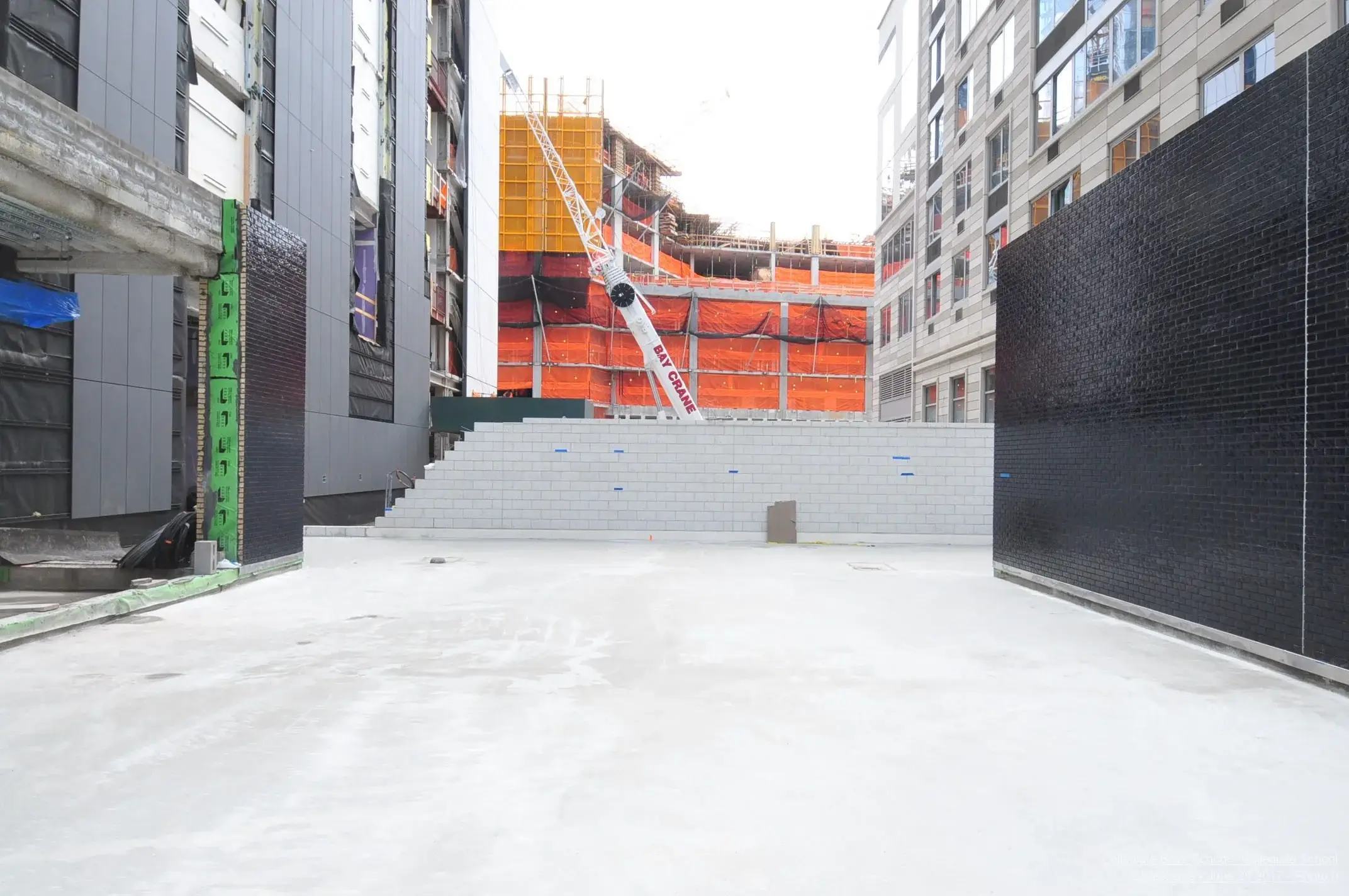 Urban construction site featuring a crane, orange safety netting, and modern buildings framing a concrete plaza.