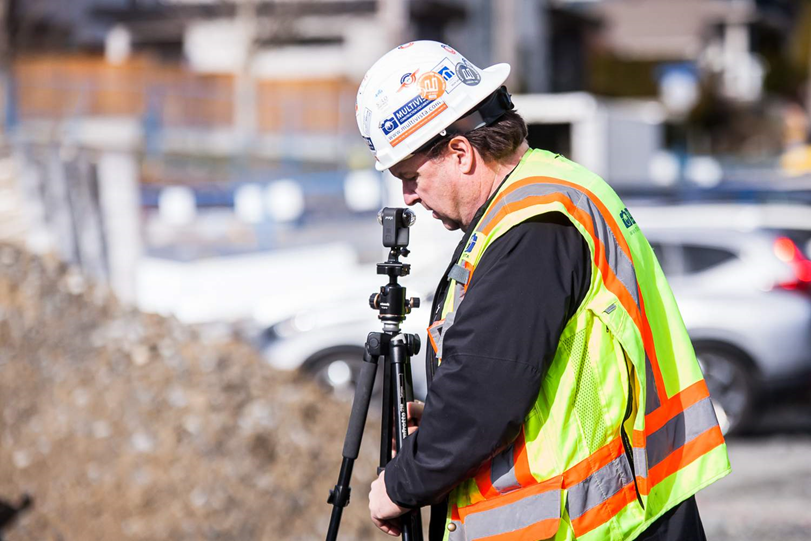 Man in hard hat and safety vest adjusting a 360 camera on a tripod at a construction site.