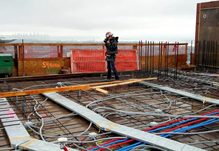 Construction site worker with hard hat surveys rebar, conduits, and cityscape from high floor.