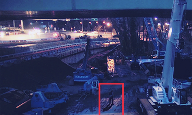 Nighttime construction site with heavy machinery, a lone worker, and illuminated traffic on a highway above.