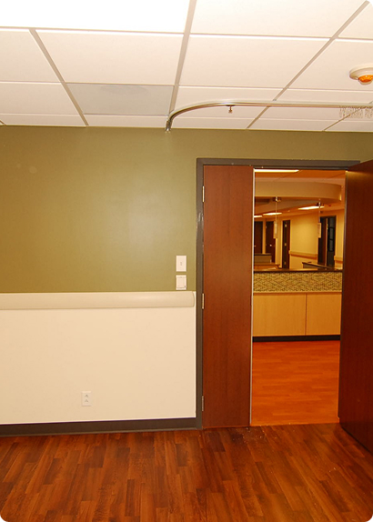 Empty patient room with olive walls, wood floor, and open door to a medical facility reception area.