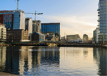 Modern city skyline with tall buildings, construction cranes, a colorful bridge over water reflecting the sky.