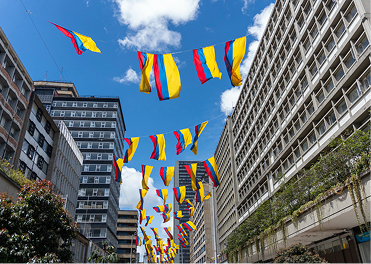 Colombian flags strung across a city street, flanked by tall buildings against a blue sky.