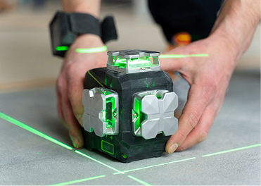 Hands adjust a black and green laser level projecting green lines on a grey floor, with a wrist device.