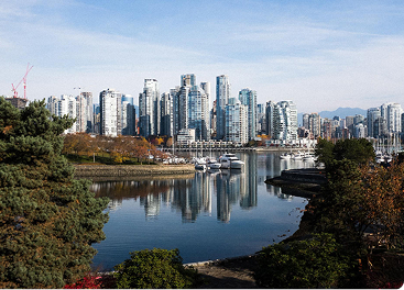 Vancouver cityscape reflects in calm water with boats, framed by green trees under a clear sky.