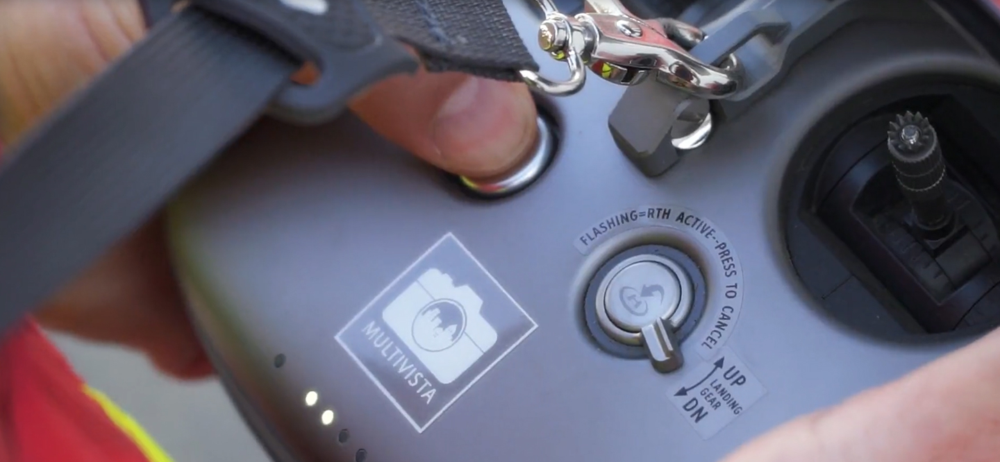 Hand pressing a button on a gray drone remote controller with Multivista logo and flight controls visible.