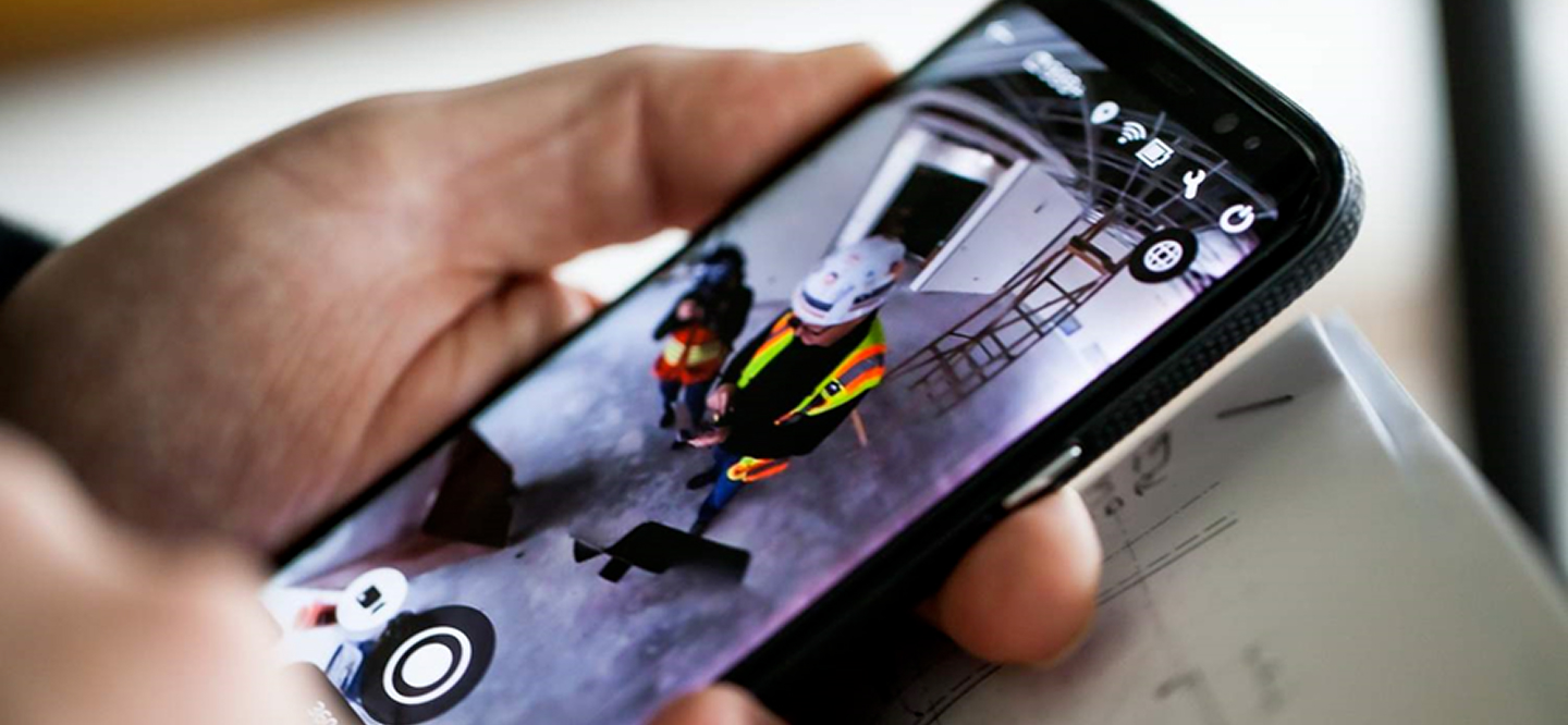 Hand holds smartphone showing a 360-degree camera view of construction workers in a building interior.