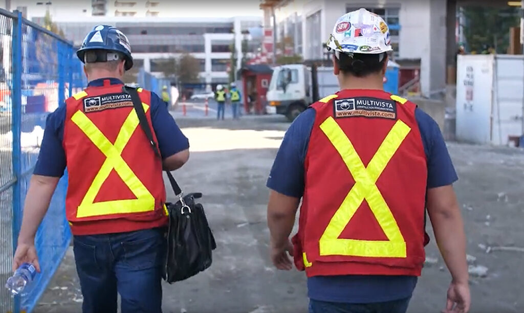 Two construction workers in Multivista safety vests and hard hats walk away from the camera on a job site.