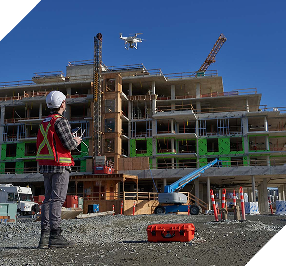 Engineer in safety vest controls a drone over a multi-story building under construction.