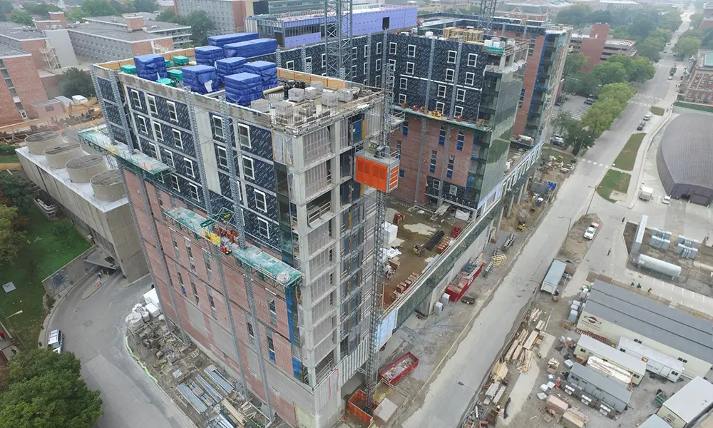 Aerial view of a large multi-story building under construction with scaffolding, an external elevator, and rooftop materials.