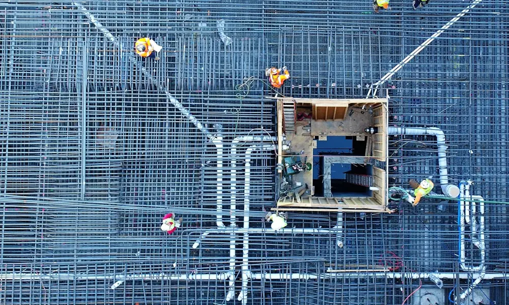 Construction site aerial showing workers on a dense rebar grid with pipes and a wooden structure.