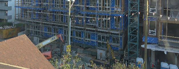 Multi-story construction site with blue scaffolding around a building and a small crane on site.