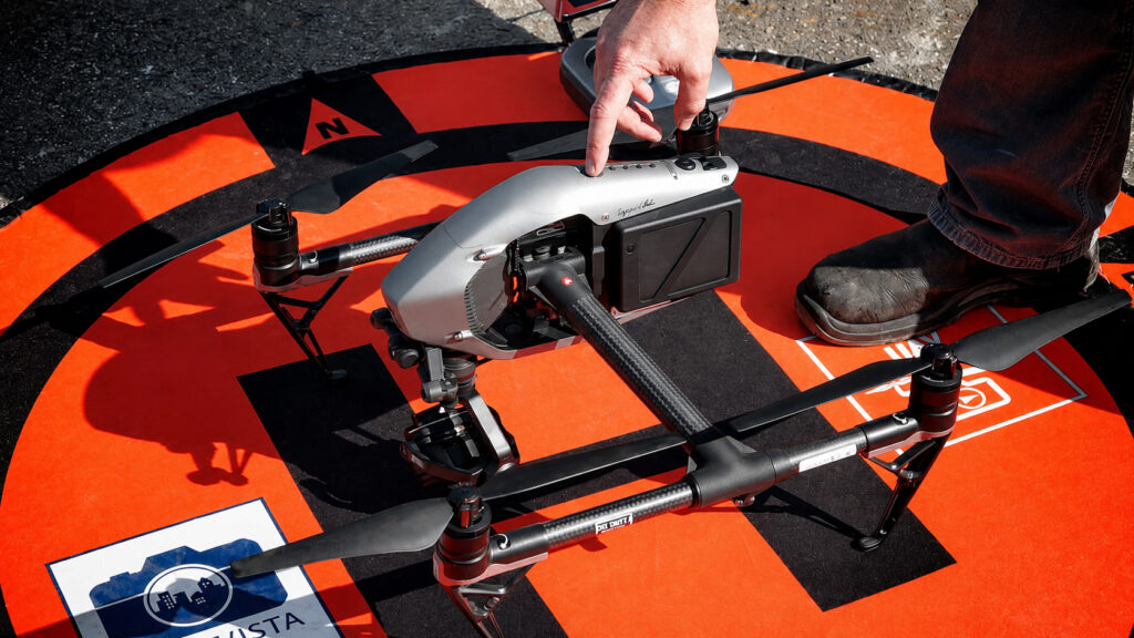 Hand presses button on a silver drone resting on an orange and black landing pad.