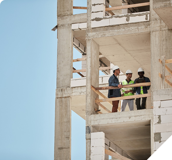 Three engineers in hard hats reviewing plans on a tablet at a concrete building construction site.