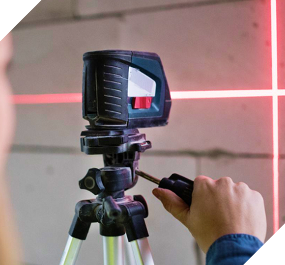 A person adjusts a laser level on a tripod, projecting red crosshairs onto a concrete wall.