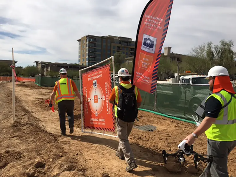 Workers in safety gear with a drone at a construction site, next to an unmanned aircraft operation sign.