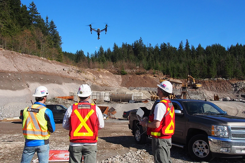 Construction workers in hard hats watch a drone flying over a job site with excavators and forest.