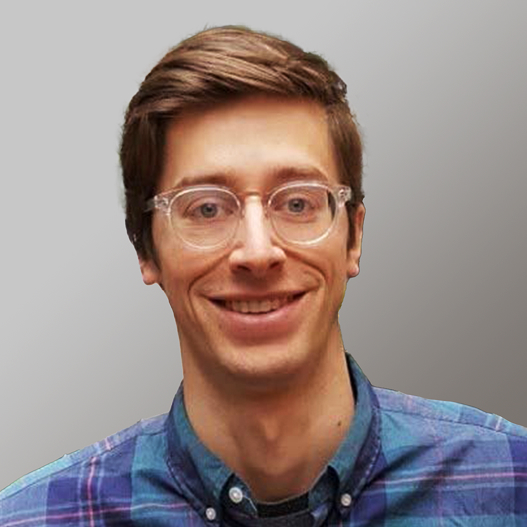 Smiling man with brown hair, clear glasses, and plaid shirt on gray background.
