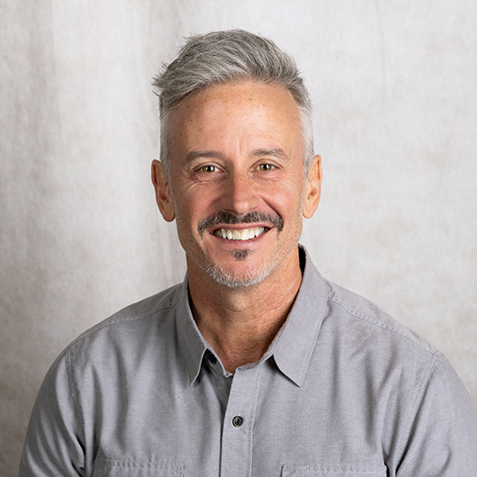 Smiling middle-aged man with gray hair wearing a gray button-down shirt, posing for a headshot against a light backdrop.