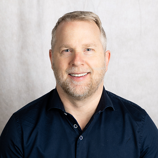 Smiling man with short gray hair and beard wearing a navy button-down shirt against a light gray background (head-and-shoulders portrait).