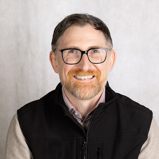Portrait of a smiling man with gray beard and glasses wearing a black zip vest, against a light neutral background.