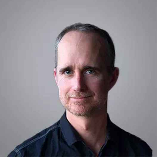 Smiling middle-aged man with graying hair and stubble in a dark collared shirt, looking at the camera.
