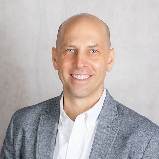 Smiling bald man in a white shirt and gray blazer, portrait against a neutral backdrop.