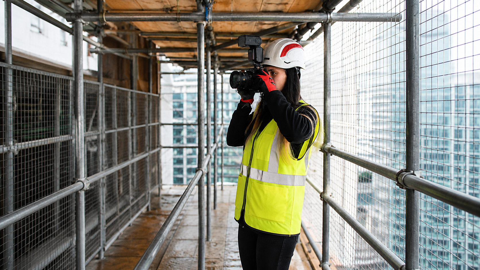 Woman in hard hat & high-vis vest photographing city skyline from scaffolding.