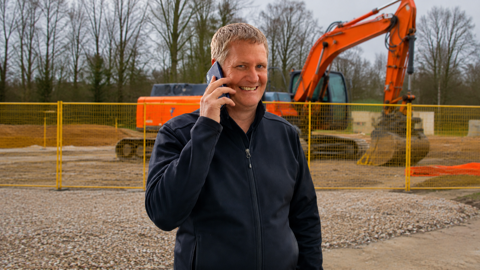 Smiling man in dark jacket talking on phone at a construction site with an orange excavator behind him.