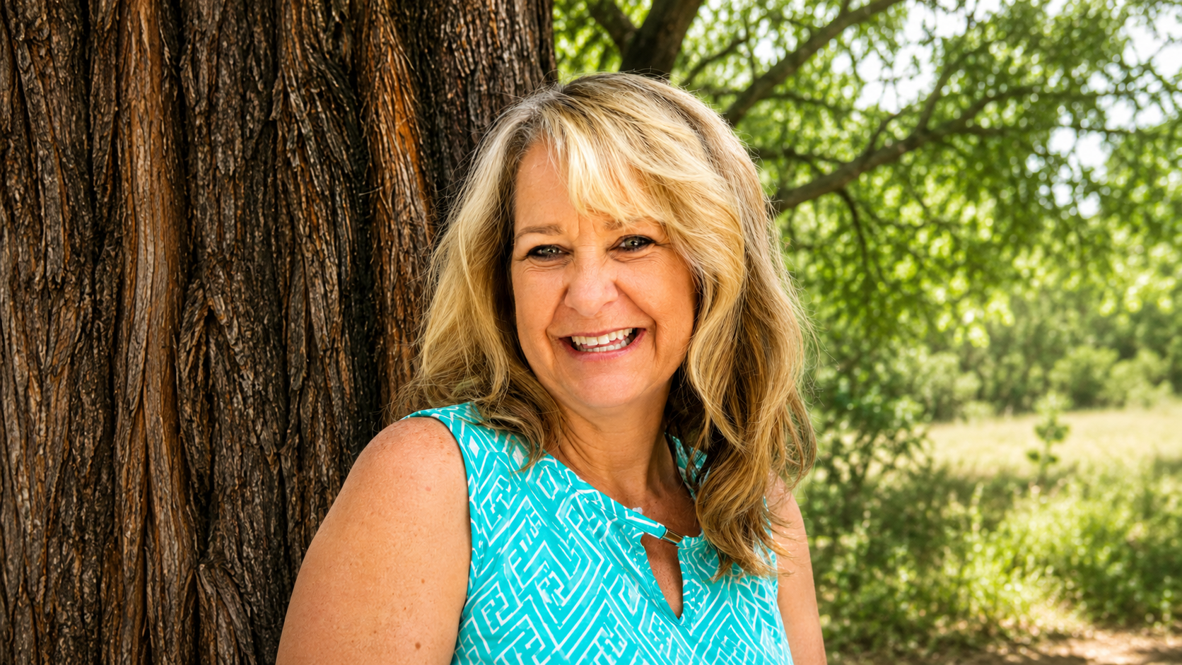 Smiling blonde woman in a turquoise patterned top leaning against a tree trunk outdoors.