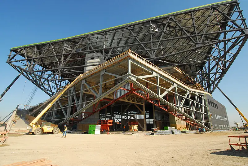 Construction of a large, modern building with a distinctive angled roof and exposed steel trusses.