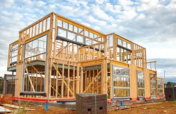 Two-story house under construction with visible wood framing and installed windows under a cloudy sky.