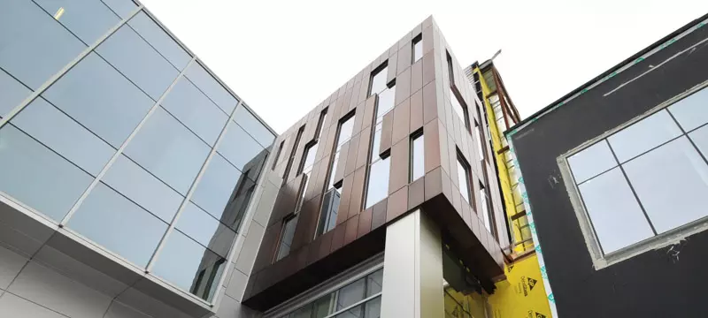 Modern building exterior featuring reflective glass, bronze panels with irregular windows, and a dark unfinished section.