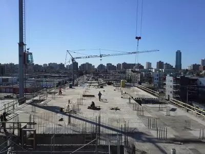 Construction site with workers, cranes, and rebar on a concrete slab, city skyline visible.